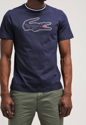 T-shirt en coton bleu marine à col rond, arborant un logo de crocodile stylisé en vert et rouge. Manches courtes et coupe ajustée.
