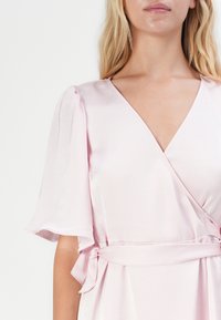 Šviesiai rožinis apsiaustas su satino audiniu, trumpomis pūstomis rankovėmis, giliu V formos iškirpimu ir surišta juosmens detalė. Sklandi tekstūra ir elegantiškas dizainas.