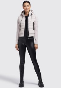Hellgraue, kurz geschnittene Puffermantel mit Kapuze, Reißverschluss und gerippten Bündchen, kombiniert mit schwarzen Leggings und Stiefeletten.