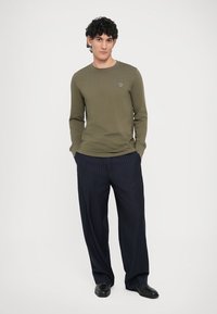 Camiseta de manga larga de color verde oliva con un pequeño logo bordado, combinada con pantalones anchos azul marino con rayas finas, completada con zapatos negros.