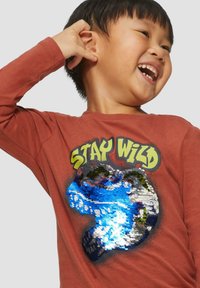 Rostfarbendes Longsleeve-Shirt mit einem Pailletten-Design, das ein blaues Wasser-Muster und den Text "STAY WILD" in Gelb zeigt.