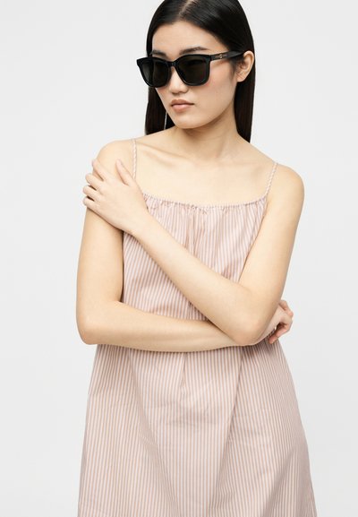 Η γυναίκα με μακριά σκούρα μαλλιά φοράει μαύρα γυαλιά ηλίου και αμάνικο φόρεμα με μπεζ και λευκές ρίγες, κρατώντας τον αριστερό της ώμο με το δεξί της χέρι.