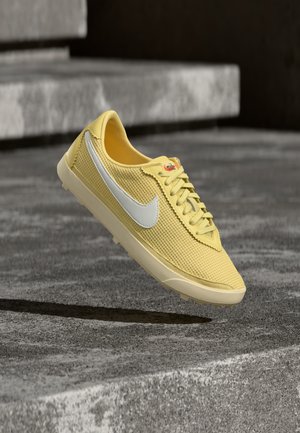 Kollane Nike toss kinga valge Swooshi logoga, tekstuurse disainiga ja paeltega ees, paigutatud hallide betoonastmete kohale.