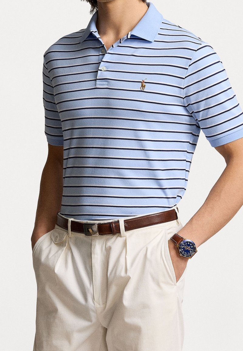 Polo bleu clair avec des rayures horizontales bleu marine, à manches courtes et col. Ceinture en cuir marron et pantalon blanc. Montre bleue au poignet.