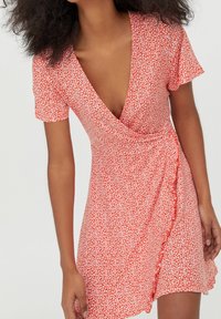 Femme portant une robe portefeuille courte rouge avec un petit imprimé floral blanc et des manches courtes, posant avec les mains près des hanches sur un fond clair.