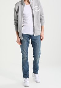 Grijze ritssweater met het logo "Tommy Jeans", witte T-shirt eronder, blauwe spijkerbroek en witte sneakers. Casual kledingensemble.