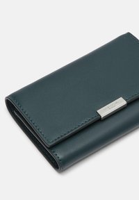 Portfel ze skóry w kolorze morskim, o gładkiej fakturze, składanym fasonie oraz srebrnym metalowym akcentem z grawerunkiem marki. Kompaktowy i minimalistyczny wygląd.