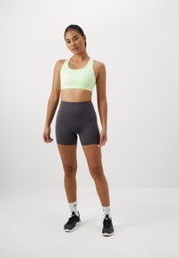 Un soutien-gorge de sport vert clair avec un col rond, associé à un short taille haute gris foncé. Le modèle porte des chaussures de sport noires et des chaussettes blanches.