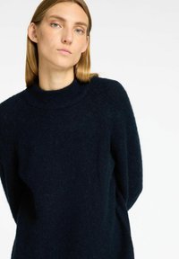 Tumši zilas krāsas džemperis, izgatavots no mīksta, teksturēta auduma, ar augstu apkakli un garām raglanu piedurknēm. Vienkāršs, brīvs dizains bez redzamām rakstzīmēm.