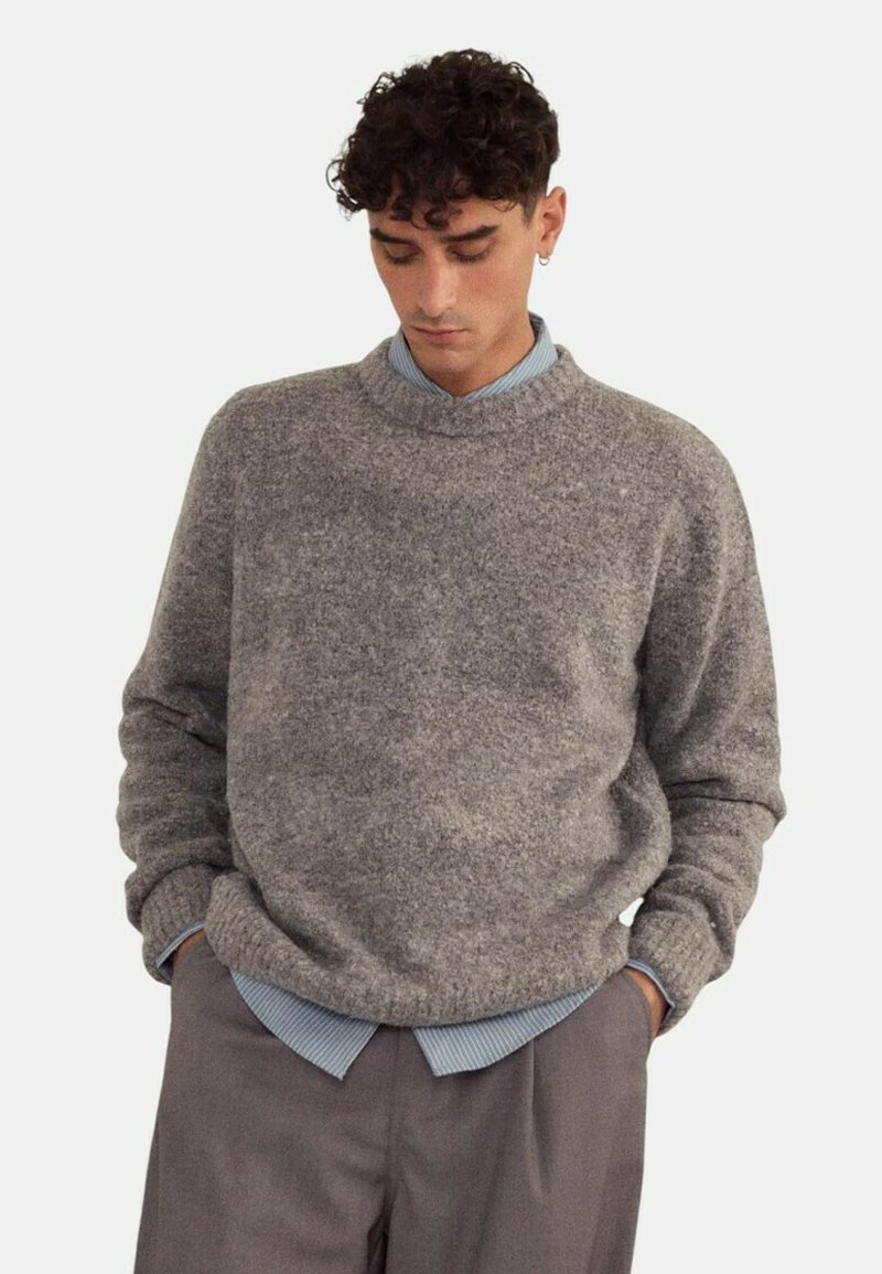 Pull en laine gris à col rond, avec une surface texturée. Porté par-dessus une chemise à col bleu clair, complété par un pantalon gris.