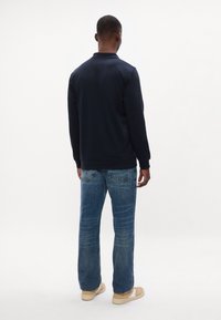 Tengerészkék pulóver magas gallérral, bordás mandzsettával, világos mosású, egyenes szárú farmerrel és krémszínű cipővel párosítva. Simogatós tapintás, hétköznapi dizájn.