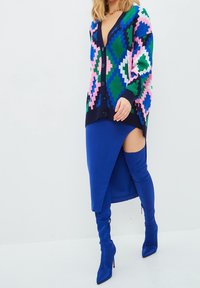 Flerfärgad stickad cardigan med ett geometriskt mönster, i kombination med en blå satinkjol med en slits på sidan och matchande knähöga stövlar.