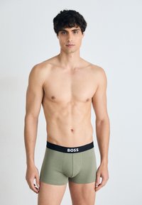 Zöld pamut keverék boxer alsó fekete elasztikus derékkal, amelyen fehér "BOSS" felirat található. A modellen nincs póló, izmos testalkatát mutatja.