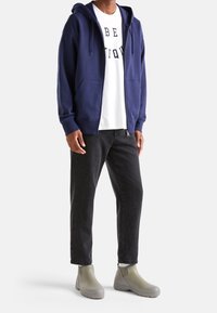 Námořnická mikina na zip vyrobená z měkké tkaniny, v kombinaci s bílým tričkem s černým textem, černými džíny a zelenými kotníkovými botami.