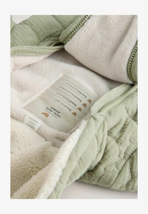 Zöld szövet kapucnis pulóver belseje puha tejszínszínű bolyhos anyaggal van bélelve; gondozási címke nyomtatott grafikával és szöveggel.