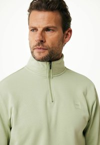 Pulóver verde claro con cuello con cremallera, tejido texturizado y un pequeño parche con el logo. El diseño presenta una silueta suave y ajustada.
