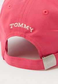 Rožinė medvilninė kepurė su baltu išsiuvinėtu užrašu "TOMMY" gale, turi metalinį uždarymą su stačiakampiu logotipo akcentu.