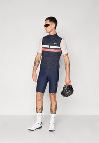 Marineblauw fietsvest met witte en rode strepen, gecombineerd met marineblauwe shorts. Model houdt een zwarte helm vast, draagt een zonnebril en witte fietsschoenen.