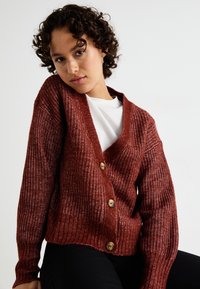 Ribbed, rust-colored cardigan featuring a deep V-neck, wooden buttons, and wide sleeves. Worn over a white shirt. Textured knit fabric.