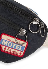 Černá látková taška s dvěma zipovými oddíly, stříbrnými doplňky a retro nášivkou v červené, modré a krémové barvě s nápisem "MOTEL Vacation."