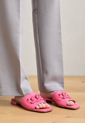 Roze natikače sa teksturiranim, pletenim detaljem na trakama, u kombinaciji sa svijetlosivim hlačama. Pozadina od drvenog poda.