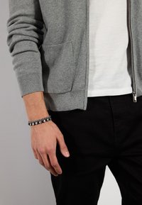 Grå glidelås cardigan med en subtil tekstur, lomme, båret over en hvit T-skjorte, parret med svarte bukser, og et sølvfarget armbånd på håndleddet.