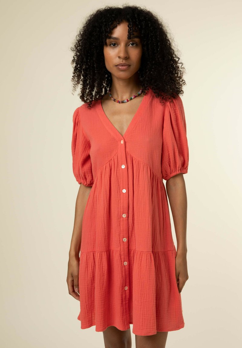 Kleid in Korallenfarbe mit V-Ausschnitt, kurzen Puffärmeln und einem gestuften Rock. Verfügt über Knopfdetails und einen strukturierten Stoff.
