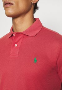 Homme portant un polo rouge avec un logo vert brodé d’un joueur de polo à cheval sur la poitrine gauche et une petite boucle d’oreille en anneau.