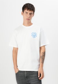 Bílý bavlněný tričko s krátkým rukávem a kulatým výstřihem. Na levé straně hrudi má modrý grafický design. Volný střih.