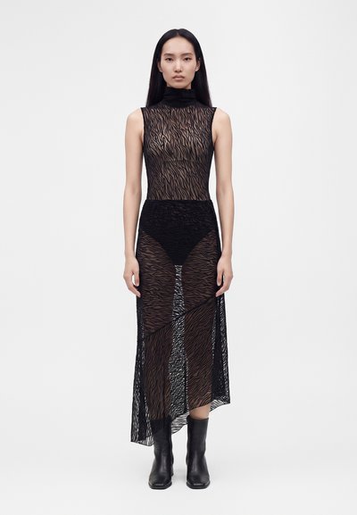 Femme debout portant une robe noire transparente sans manches à imprimé zèbre sur des sous-vêtements noirs et des bottines noires, devant un fond uni.