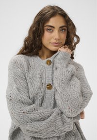 Graue Strickjacke mit strukturiertem Muster, ausgestattet mit drei auffälligen goldenen Knöpfen entlang der Vorderseite und weiten, übergroßen Ärmeln.