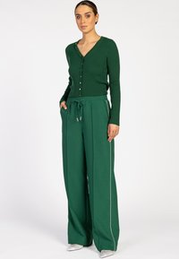 Vrouw in een donkergroene geribbelde knoopcardigan en wijde groene broek met zijstrepen, staande met handen in de zakken.