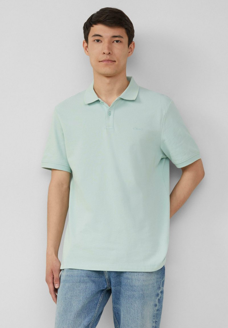 Lichtgroene polo met een klassieke kraag, korte mouwen en subtiele textuur. G gedragen met lichtblauwe jeans. Eenvoudig logo op de borst.