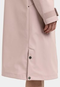 Lys lyserød regnfrakkeærme med velcrobånd og hånd ved jakkeafsætning med to sorte trykknapper på sidelommerne.