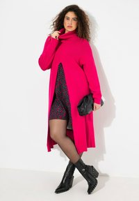 Un cardigan rose à manches longues avec un col montant, associé à une robe noire à motifs. Des bottines noires et une pochette noire complètent le look.