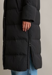Hand rustend op een zwart gewatteerd jack met rits, gedragen over blauwe jeans tegen een effen achtergrond.