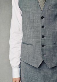 Chaleco de tela gris con un patrón texturizado, que presenta cinco botones negros, dos bolsillos y un dobladillo puntiagudo, superpuesto sobre una camisa blanca.