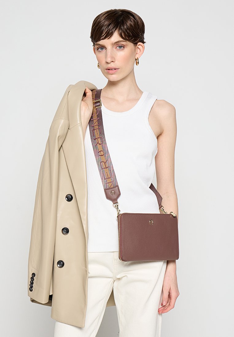 Braune, strukturiertes Lederhandtasche mit Reißverschluss und abnehmbarem Tragegurt mit einem mehrfarbigen Muster. Das Model trägt ein weißes Tanktop und einen beigen Mantel.