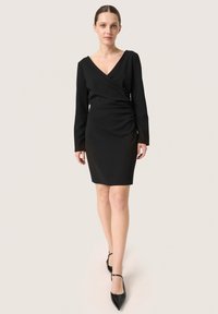 Schwarzes Wickelkleid mit langen Ärmeln, V-Ausschnitt und gerafftem Seitendetail, kombiniert mit schwarzen Pumps mit spitzer Zehenpartie. Glatte Stofftextur.
