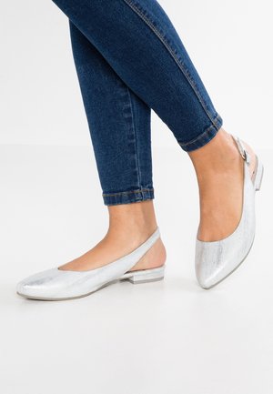 Fødder iført sølvfarvede slingback-flats kombineret med mørkeblå skinny jeans mod en ensfarvet lys baggrund.