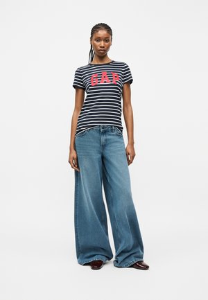 Kvinne iført svart og hvit stripete GAP-t-skjorte og vide blå jeans, stående foran en ensfarget hvit bakgrunn.