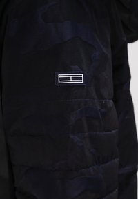 Chaqueta de plumas de camuflaje navy con costuras horizontales, que cuenta con un parche de logo blanco en la manga y una textura suave y ligera.