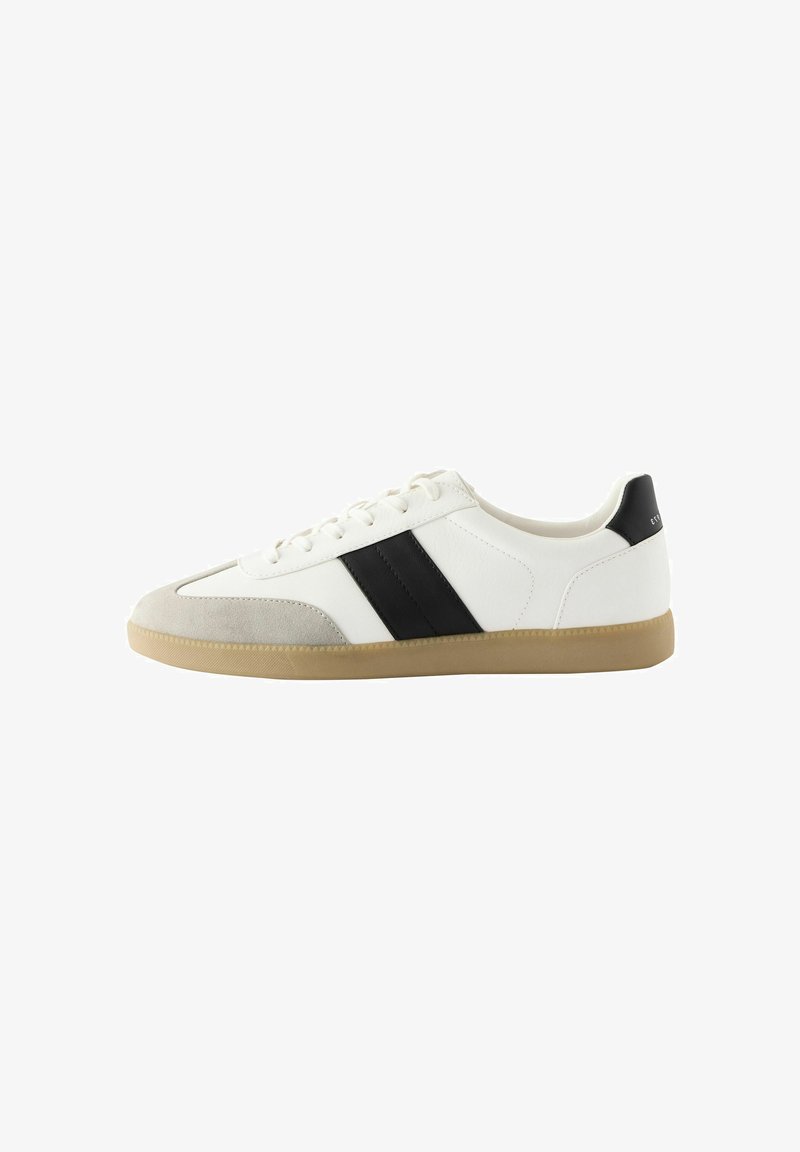 Sneaker blanc en cuir et daim, avec des rayures horizontales noires, une semelle en caoutchouc beige et des lacets plats.