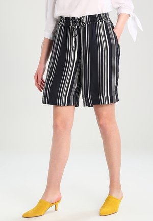 Femme portant un short rayé vertical noir et blanc avec une taille élastique et une blouse blanche avec des manches nouées.