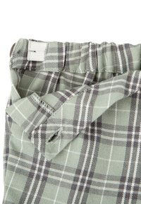 La tela a cuadros verde presenta patrones de cuadrícula en negro y blanco. Cintura elástica con una etiqueta blanca y detalles de costura visibles. Textura suave.