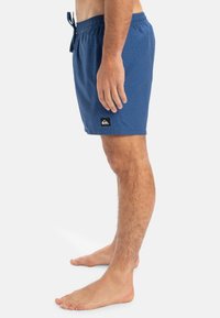 Modré plavky vyrobené z textúrovanej látky, so elastickým pásom a šnúrkou na stiahnutie, a čiernou nášivkou loga značky.