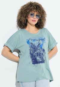 Camiseta oversized en verde claro con mangas cortas enrolladas, que presenta una ilustración de un tranvía azul y el texto "Portugal" en una fuente de estilo casual.