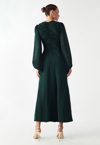 Dlhé, tmavozelené šaty z hladkej látky, s vypučanými dlhými rukávmi, zbieraním na chrbte, dĺžkou po členky a so zipsovým uzáverom.