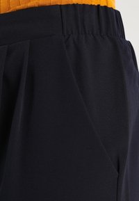 Pantaloni blu navy con tessuto testurizzato, dotati di una ampia cintura elastica e leggere pieghe, che mostrano una vestibilità rilassata e una superficie liscia.