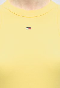 Záber na osobu nosiacu žlté tričko s okrúhlym výstrihom a malým vyšitým logom vlajky v červenej, bielej a modrej farbe v strede hrudníka.
